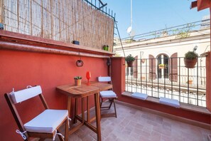 A balcony with a wooden table and two chairs, a red wall, and a view of a building with balconies. - A cozy 25-square-meter studio apartment, ideal for two people, located on the fourth floor of a building (no elevator) in the central Rione Monti district, just steps away from the Imperial Forums. The apartment enjoys a prime location, very close to the (Rome)