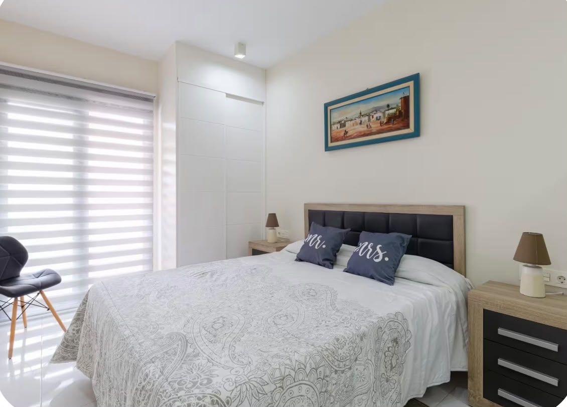 A bedroom with a bed, two bedside tables, a chair, a painting on the wall, and a window with blinds.