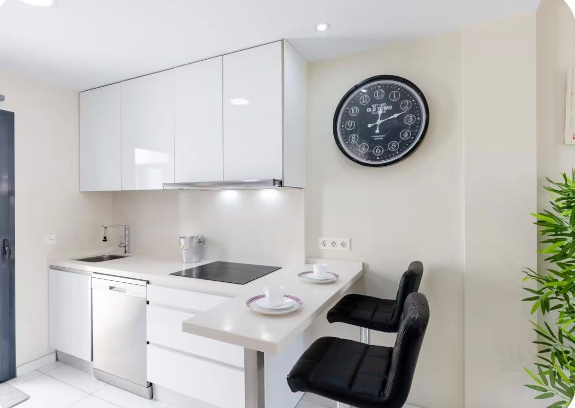 Une cuisine moderne avec des meubles blancs, une horloge noire au mur et un petit coin repas avec une chaise noire.