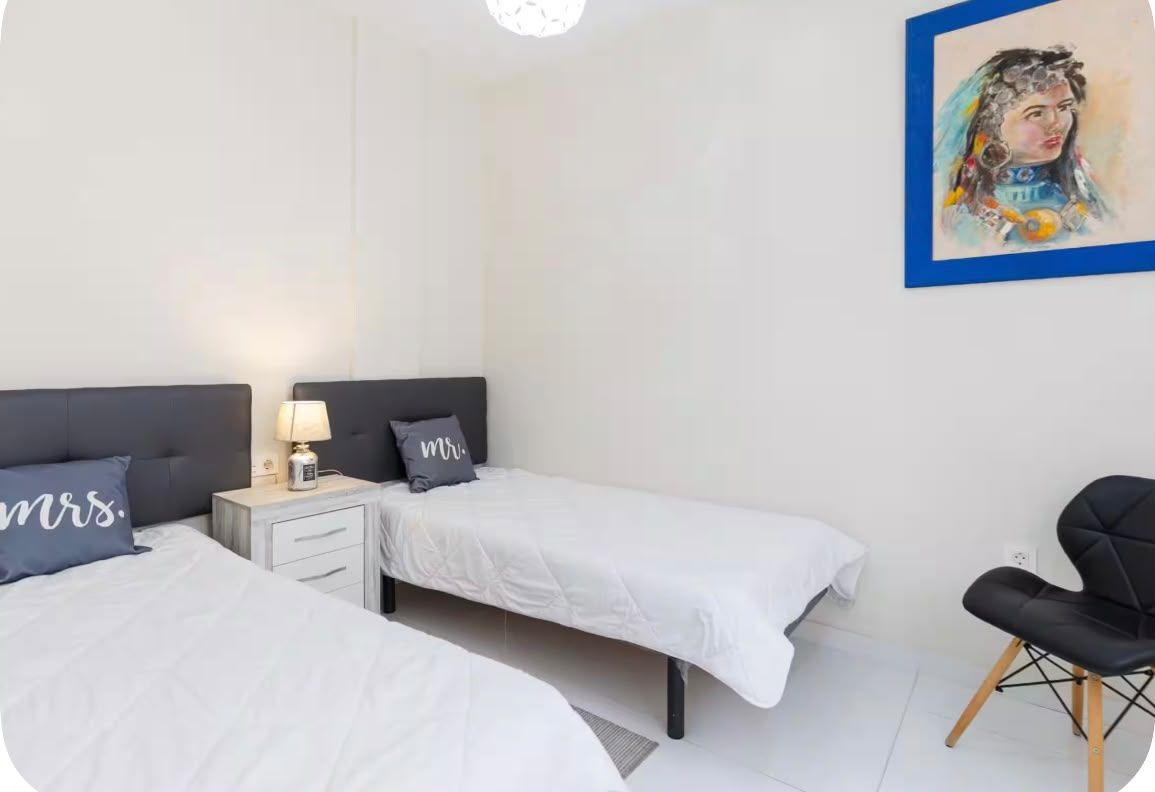 Une chambre d’hôtel avec deux lits simples, une table de chevet avec une lampe, une chaise et un tableau au mur.