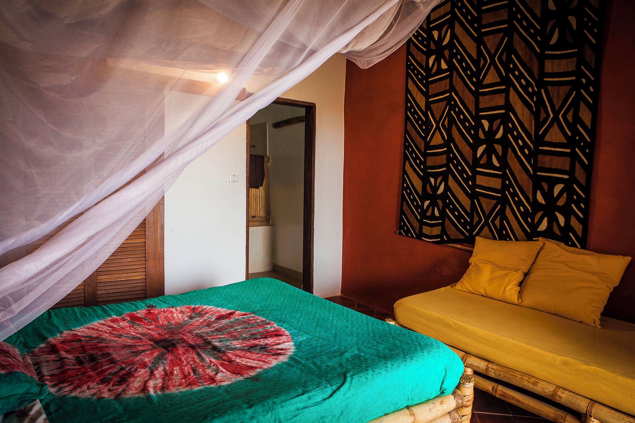 Una habitación con una cama cubierta por una manta de diseño verde y rojo, un sofá amarillo con un cojín a juego y un panel decorativo en la pared.