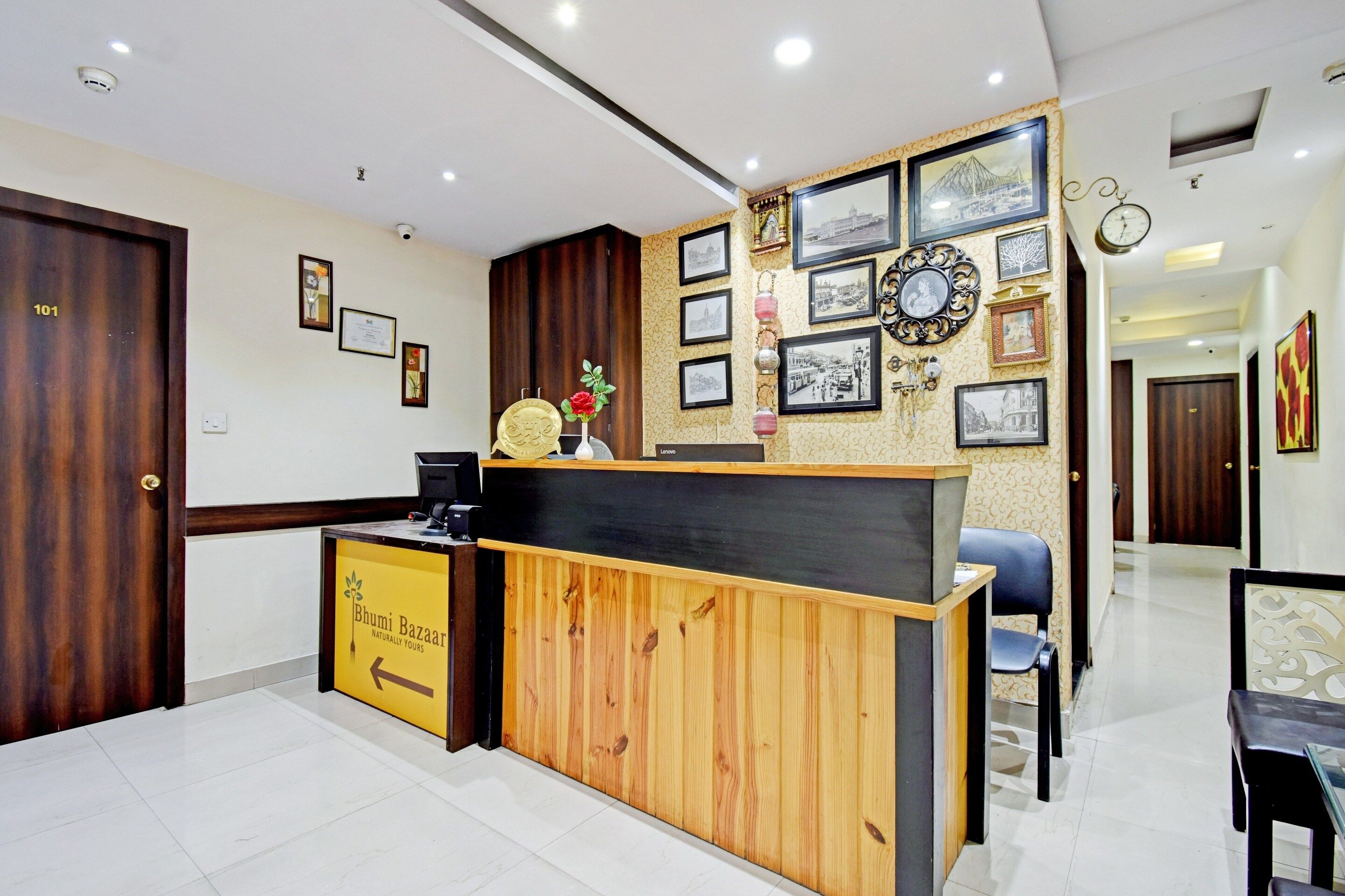 Un mostrador de recepción con detalles de madera, una barra negra y un cartel para Bhumi Bazaar. Hay cuadros enmarcados y un reloj en la pared.