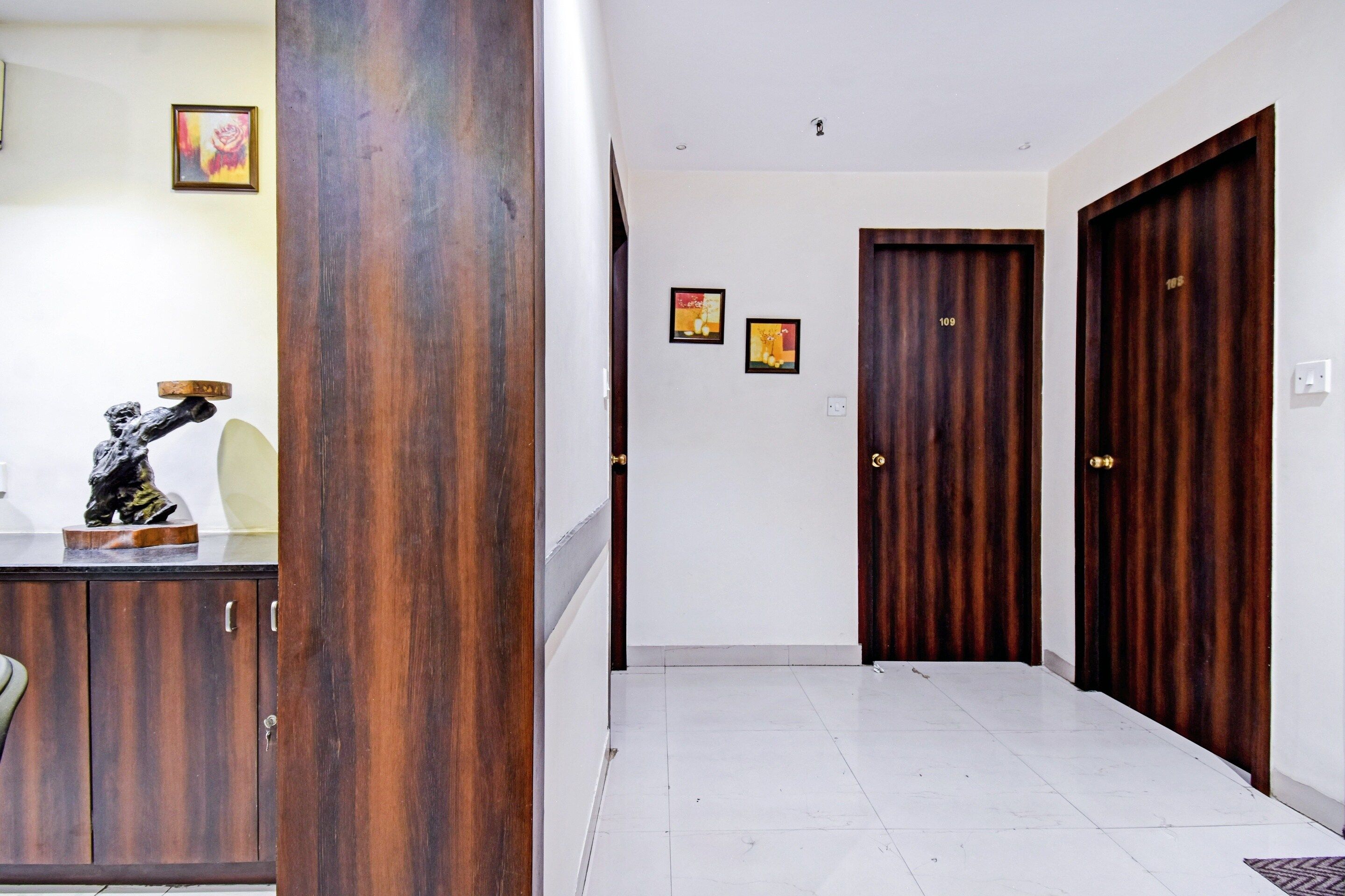 Un pasillo con una puerta de madera, una pared blanca con dos cuadros enmarcados y una estatua sobre un mueble.