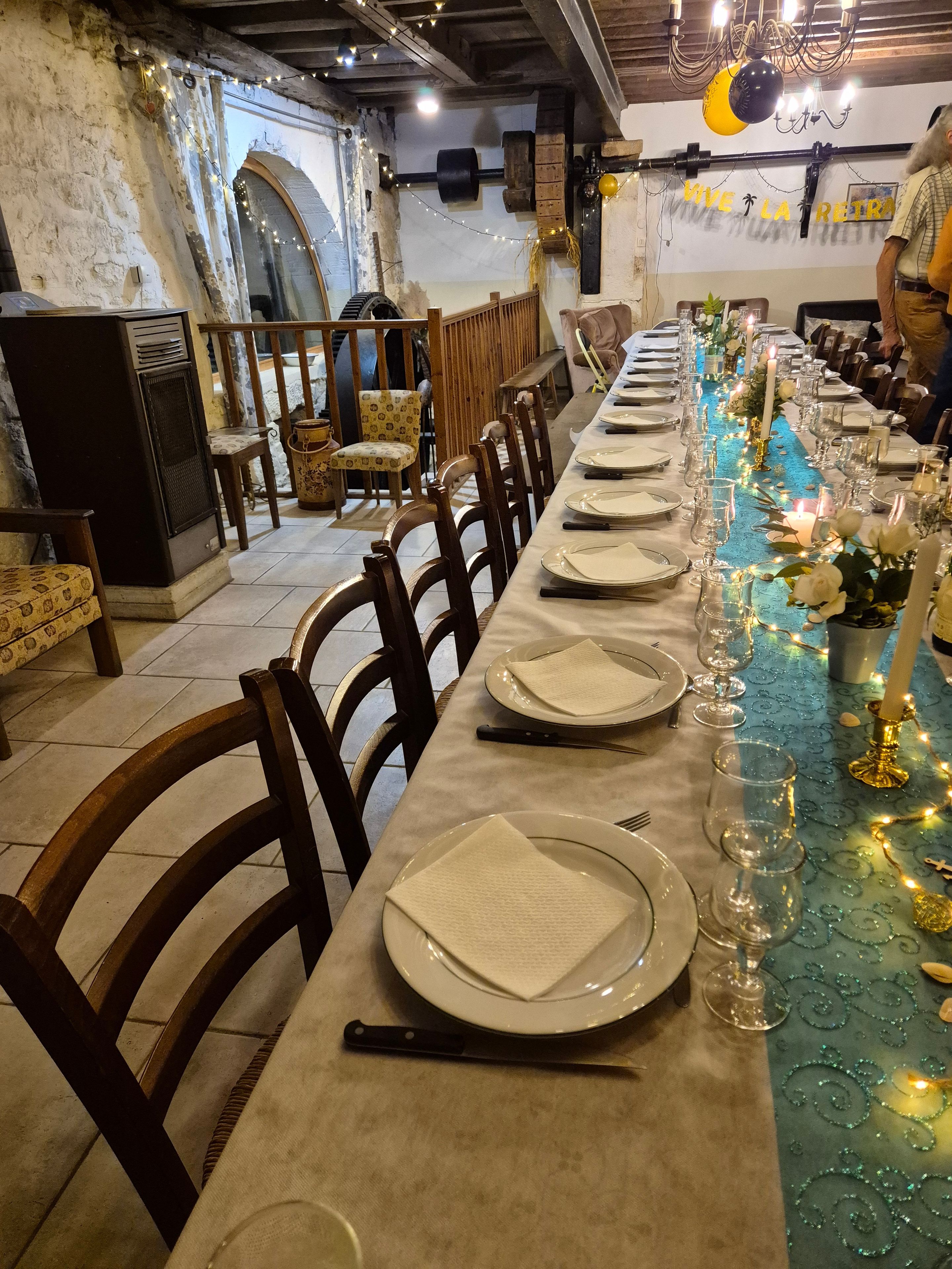 Une longue table à manger, dressée avec des assiettes, des couverts et des verres, et décorée de guirlandes lumineuses et de fleurs.