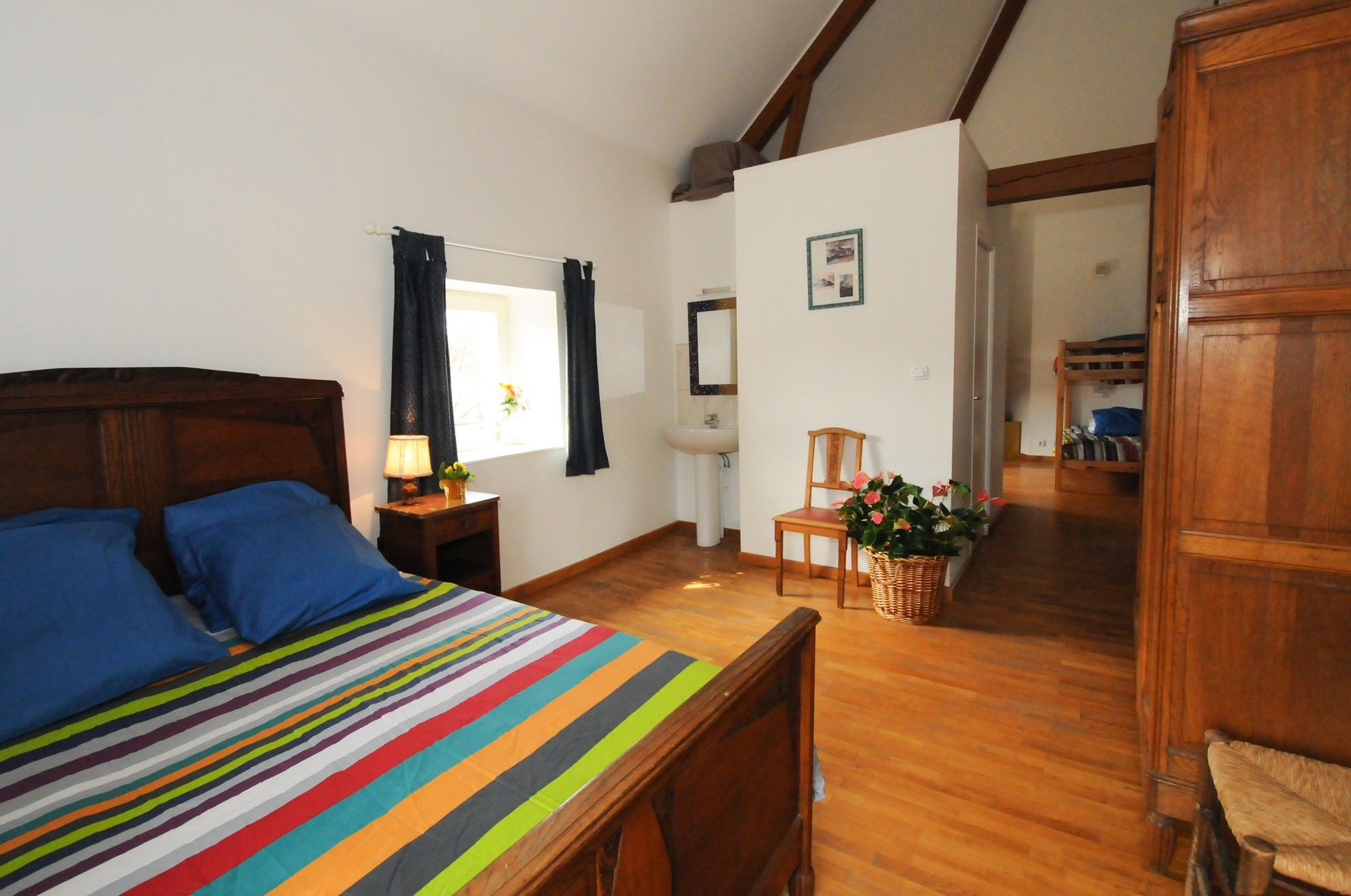 Una camera da letto con un letto in legno, un comodino con una lampada, una sedia, una finestra con tende, un lavandino e un armadio in legno.