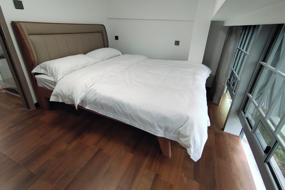 A modern bedroom with a wooden bed frame, white bedding, and a large window providing a view of the outdoors.