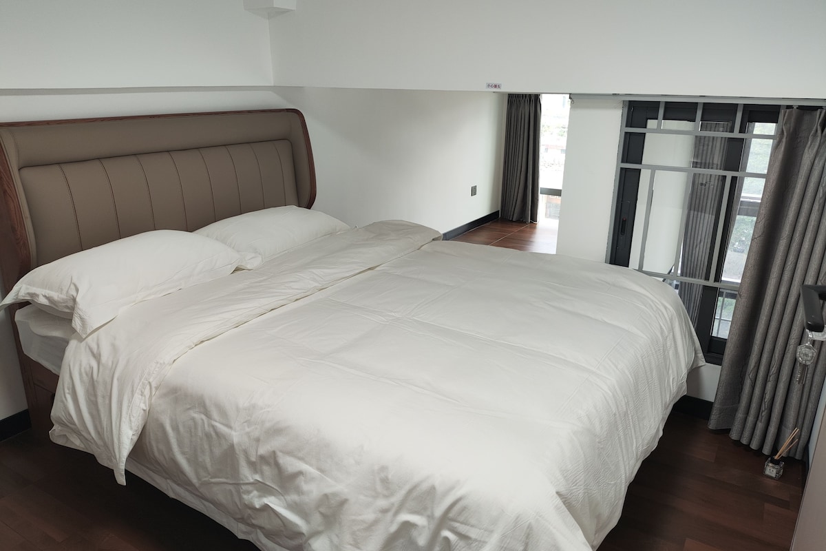 A hotel room with a large bed, a headboard, a window with curtains, and a wooden floor.
