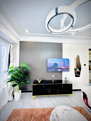 A modern living room with a flat-screen TV mounted on a wall, a black entertainment unit, a large potted plant, and a marble coffee table. - Elagant 2BR in lavington/kilimani/kileleshwa w/pool gym near malls central place (Nairobi)