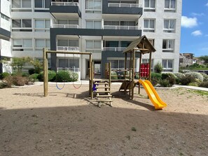 A playground with swings and a slide, surrounded by a sandy area with some greenery. - Vive La Serena con comodidad y estilo. Departamento nuevo completamente equipado (La Serena)