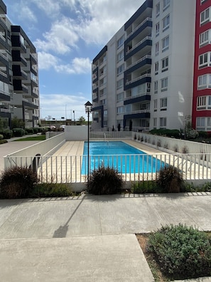 Ein Schwimmbecken, umgeben von einem weißen Zaun, mit mehrstöckigen Gebäuden im Hintergrund.