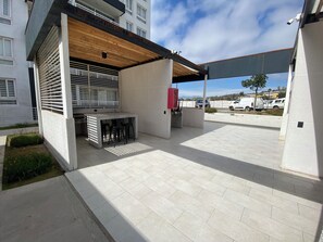 An outdoor patio area with a concrete floor, a wooden pergola, a small bar area with stools, and a red electrical box. - Vive La Serena con comodidad y estilo. Departamento nuevo completamente equipado (La Serena)