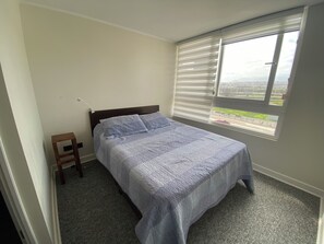 Ein Schlafzimmer mit einem Bett, einem Fenster mit Jalousien, einem kleinen hölzernen Hocker und einem Teppichboden.