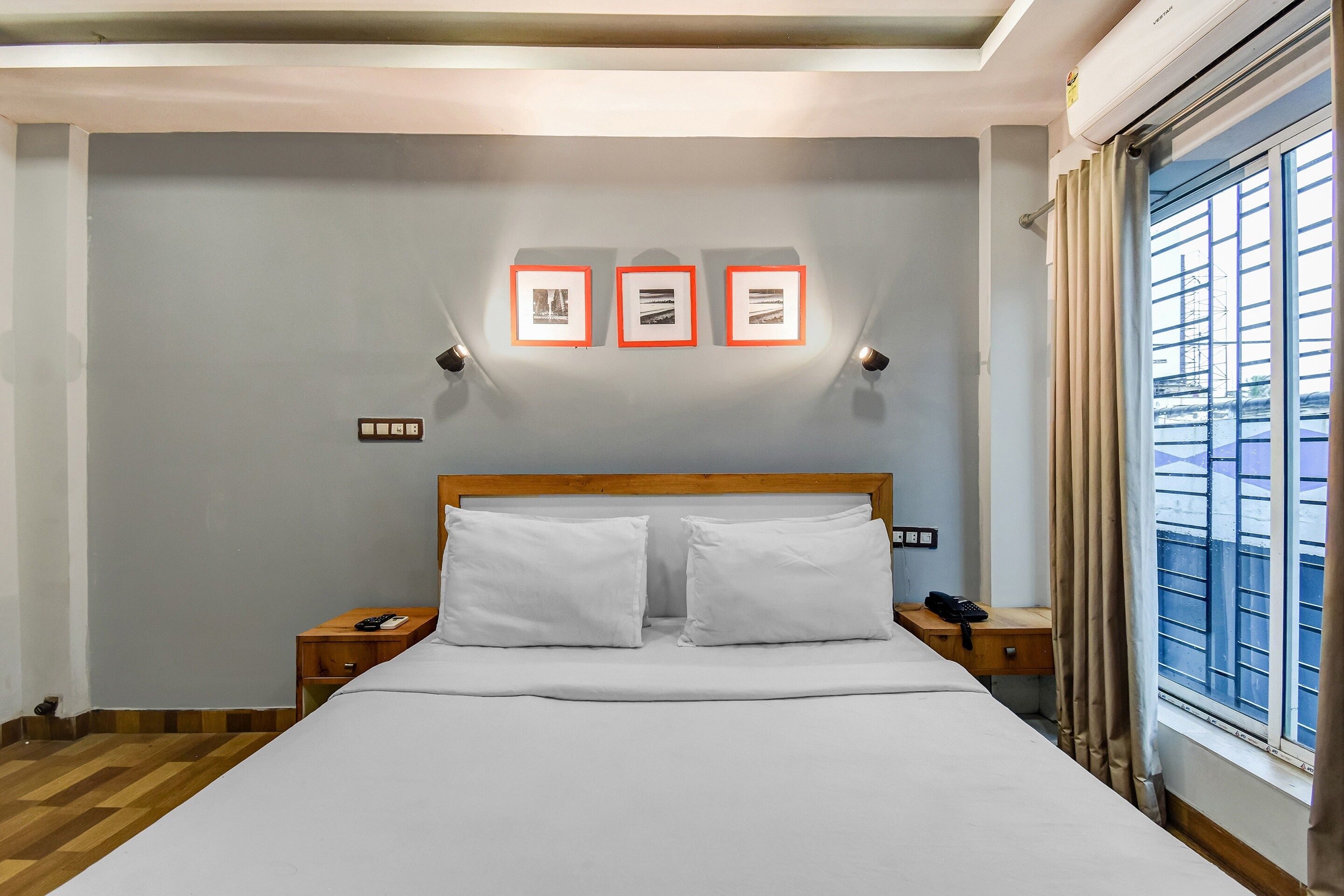 Een hotelkamer met een bed, twee nachtkastjes, een raam met gordijnen en drie ingelijste foto's aan de muur.