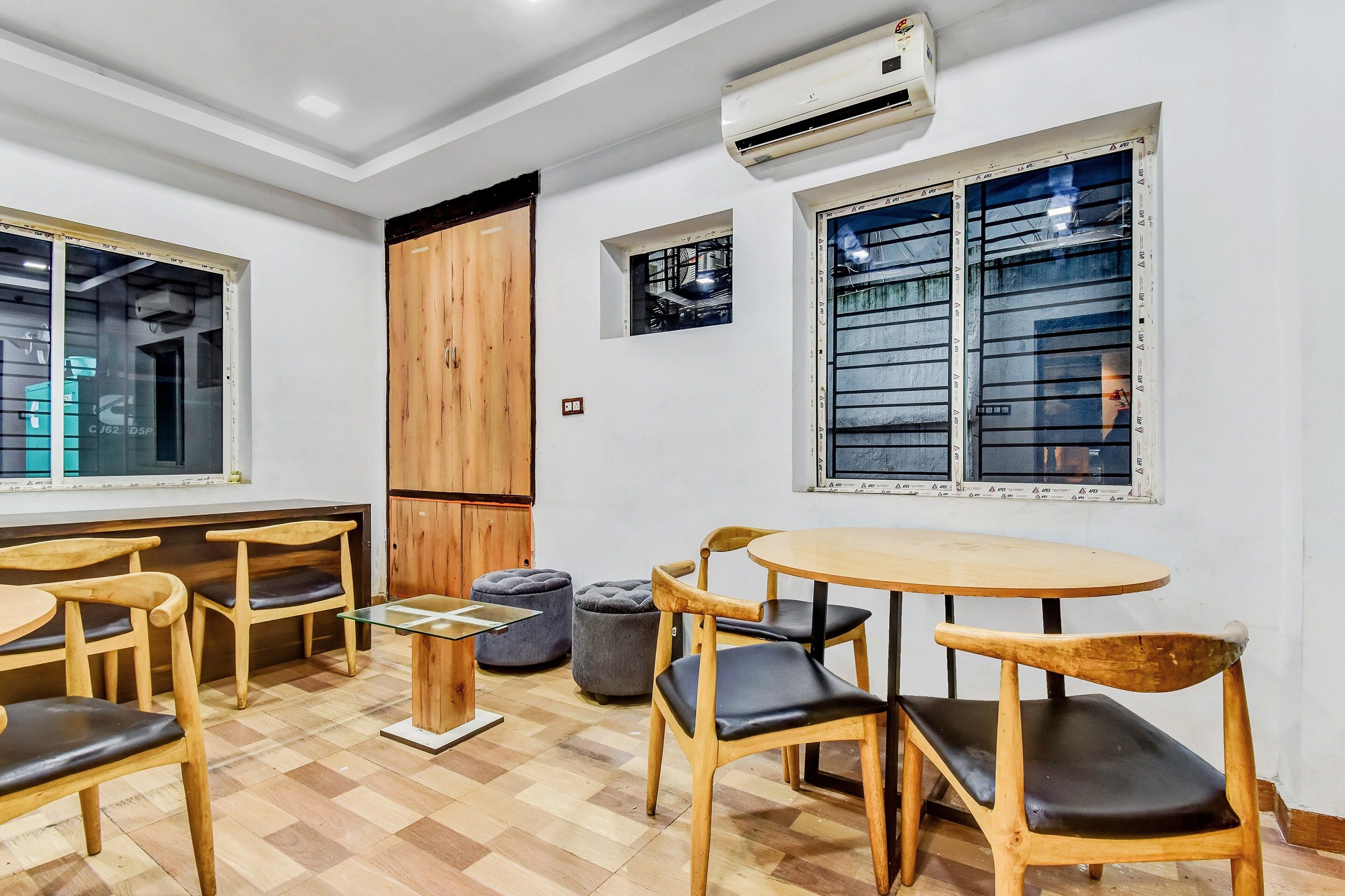 Una habitación con piso de madera, una mesa redonda y varias sillas. Hay un equipo de aire acondicionado en la pared y una ventana con persianas.