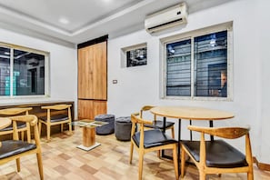 Een kamer met een houten vloer, een ronde tafel en meerdere stoelen. Er is een airconditioning op de muur en een raam met zonwering.