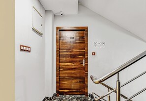 A hallway with a wooden door labeled '201', a wall-mounted light switch, and a metal staircase railing. - Hotel O MB International (Kolkata)