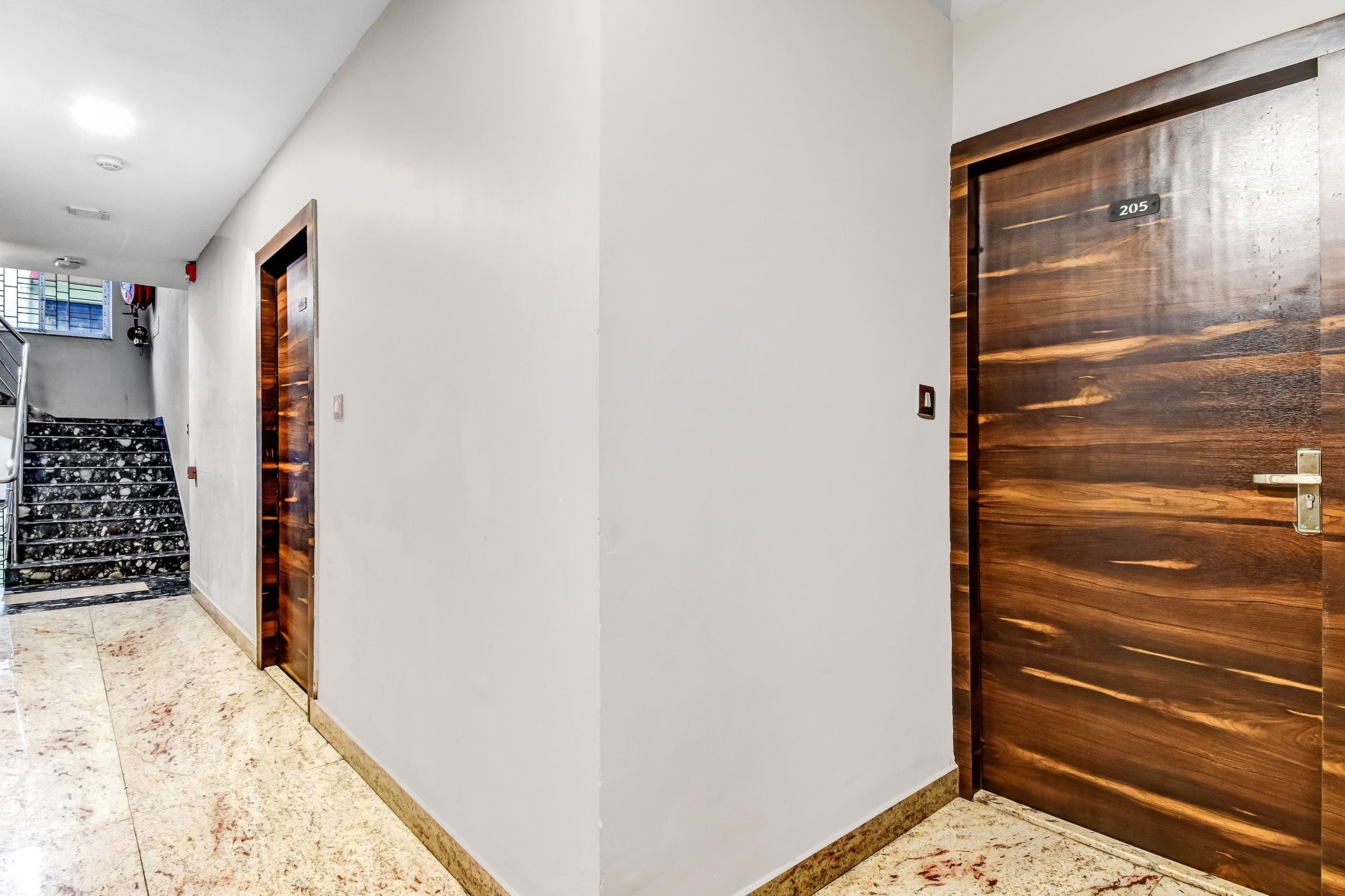Un couloir avec deux portes en bois, un escalier recouvert de carreaux noirs et blancs, et un mur blanc sur lequel est fixée une télévision.