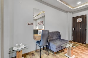 Habitación de hotel con sofá, una silla, una mesita con superficie de vidrio, un espejo y una puerta de madera.
