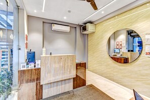 A reception area with a wooden reception desk, a mounted air conditioner, a round mirror on the wall, and a glass partition with a view of the outside. - Hotel O MB International (Kolkata)