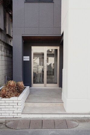A building entrance with a glass door, a small garden area with plants, and a sign on the wall. - ISSHUKU Toshima ikebukuro3 (Toshima-ku)