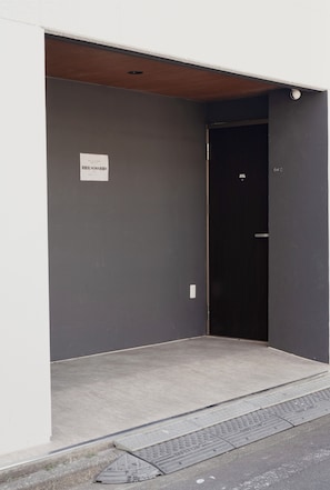 A narrow entrance to a building with a dark grey wall and a white door. A sign with Chinese characters is visible on the wall. - ISSHUKU Toshima ikebukuro3 (Toshima-ku)