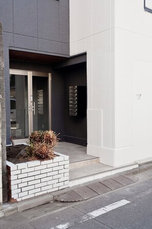 A modern building entrance with a glass door, a small garden area with plants, and a metal gate. - ISSHUKU Toshima ikebukuro3 (Toshima-ku)