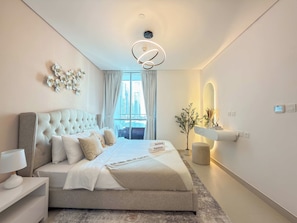 A modern bedroom with a tufted headboard, a large bed with multiple pillows, a bedside table with a lamp, a decorative wall piece, and a view of the cityscape through the window. - Rentico | 1BR Marina Waterfront | Pool & Cinema (Dubai)