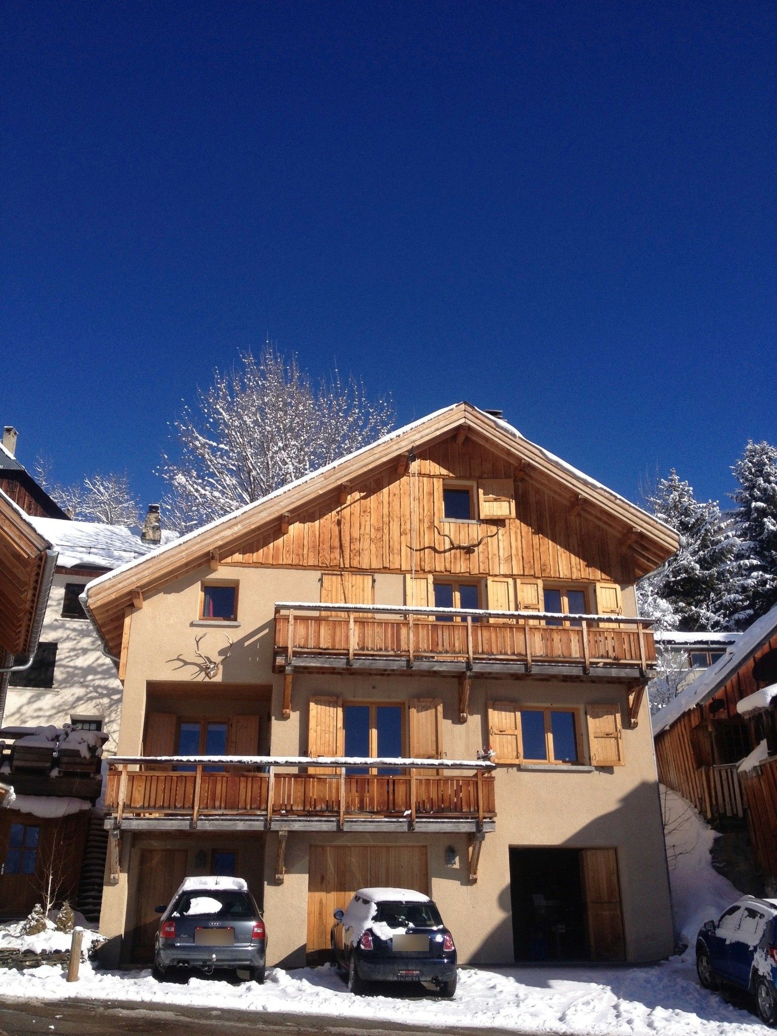 A two-story wooden chalet with a balcony, a garage with two cars, and a clear blue sky.