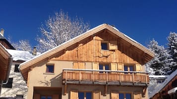 A two-story wooden chalet with a balcony, a garage with two cars, and a clear blue sky.