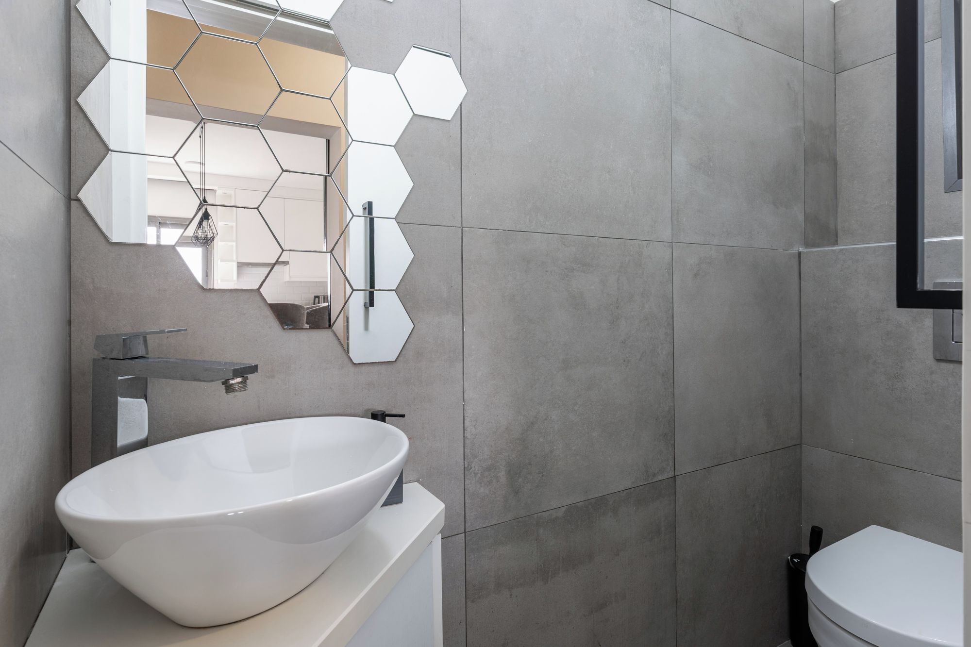 A modern bathroom with a large white vessel sink, a hexagonal mirror, and a toilet.