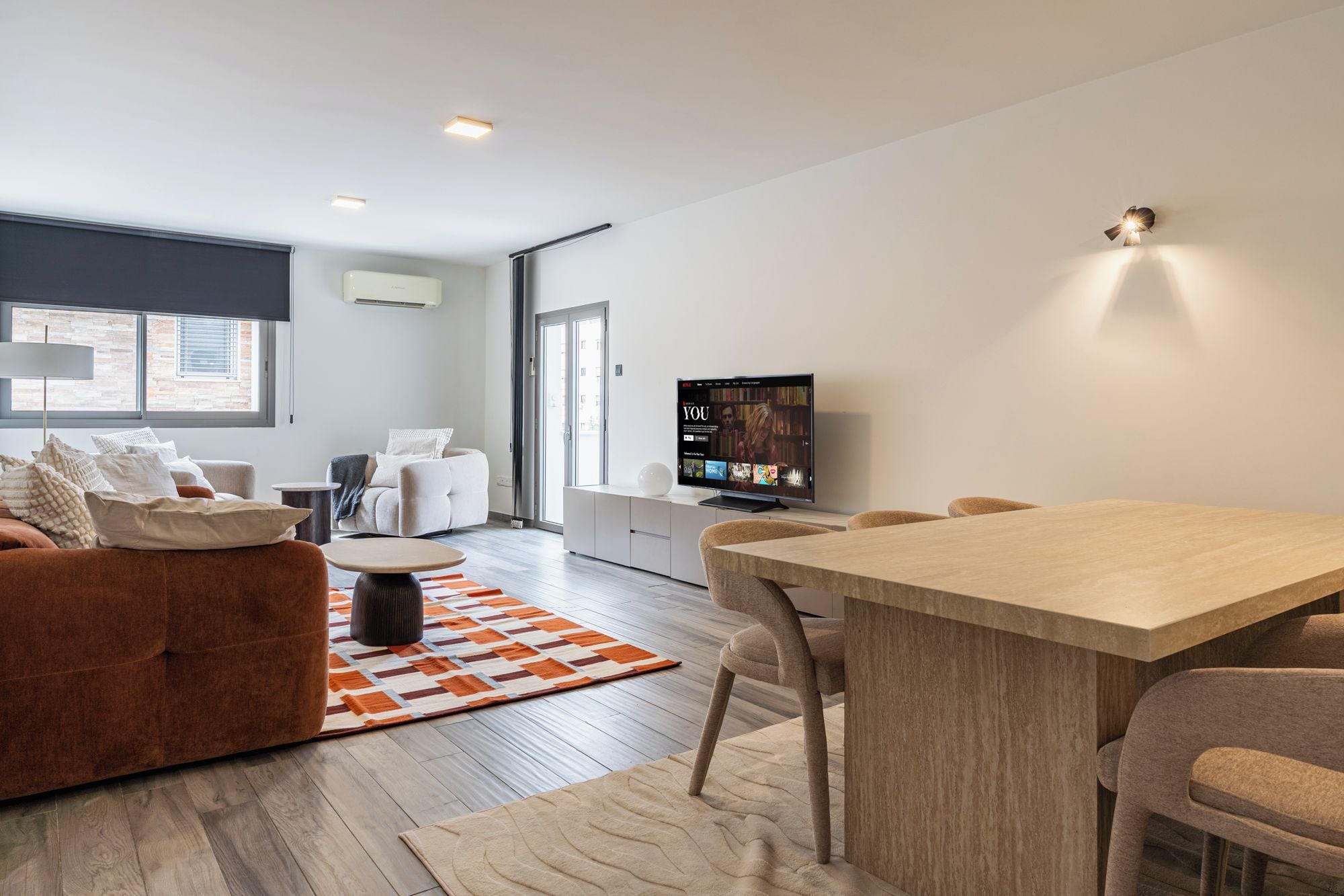 A modern living room with a sofa, a coffee table, and a TV. There is a dining area with a table and chairs.
