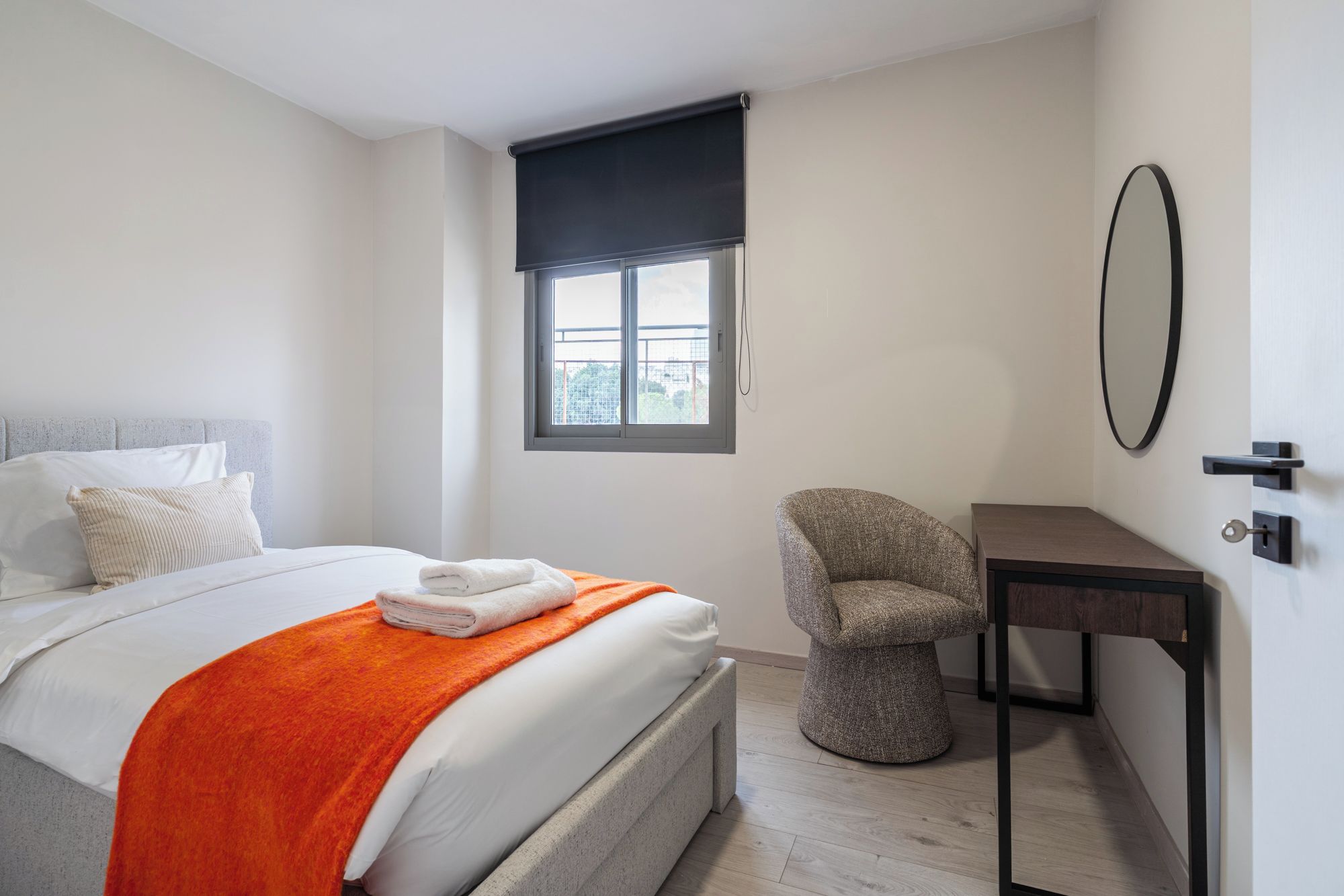A bedroom with a bed, a chair, a desk, a window with a blind, and a door with a key.