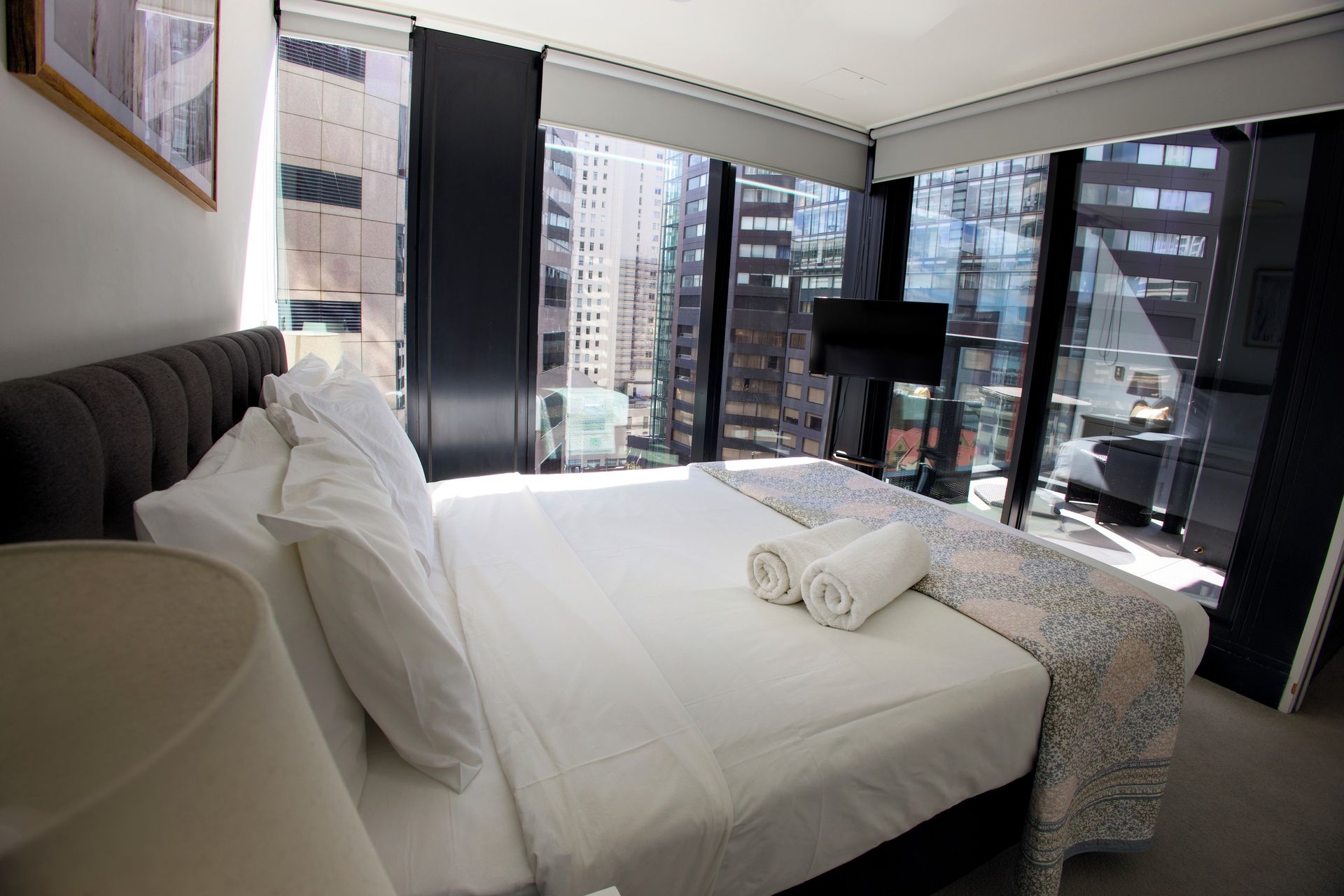 Une chambre d’hôtel dotée d’un grand lit, offrant une vue sur le paysage urbain grâce à de larges fenêtres du sol au plafond, d’un téléviseur à écran plat et d’un design moderne.