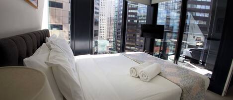 A hotel room with a large bed, a view of the cityscape through floor-to-ceiling windows, a flat-screen TV, and a modern design.