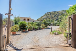 A rural entrance with a gravel path leading to a house with a tiled roof, surrounded by greenery and a hill in the background. - Villa Santuiri Can Tomeu - By Dream Villas Pollensa (Pollensa)