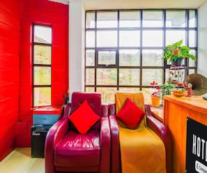 Une chambre d’hôtel avec un mur rouge, une grande fenêtre, un fauteuil en cuir, un bureau en bois et une enseigne sur laquelle on peut lire « HÔTEL ».