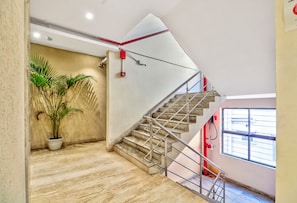 Una escalera con barandilla de metal, una planta en maceta y un detector de incendios rojo en la pared.