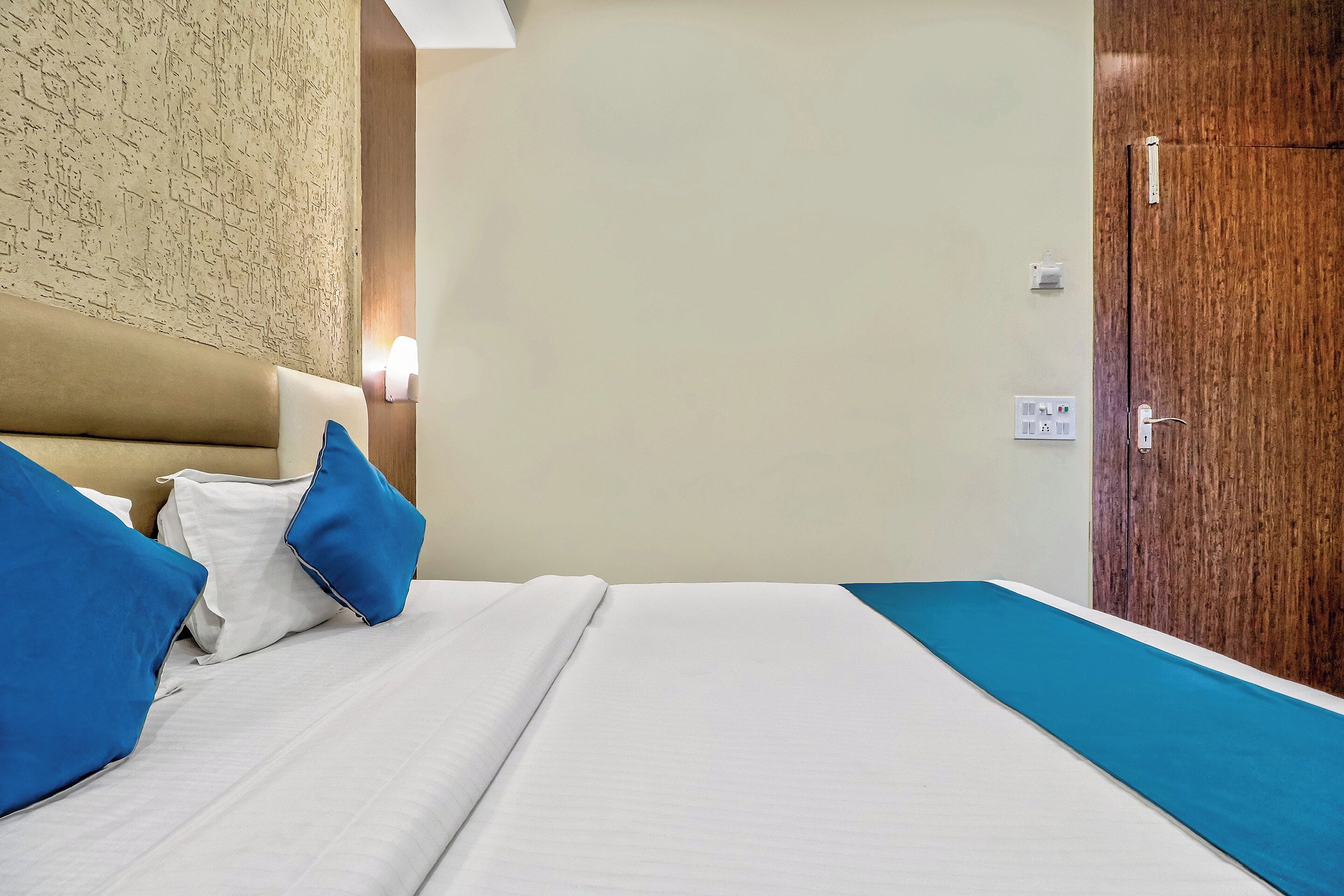 Habitación de hotel con cama, almohadas, mesita de noche, lámpara de pared y puerta de madera.