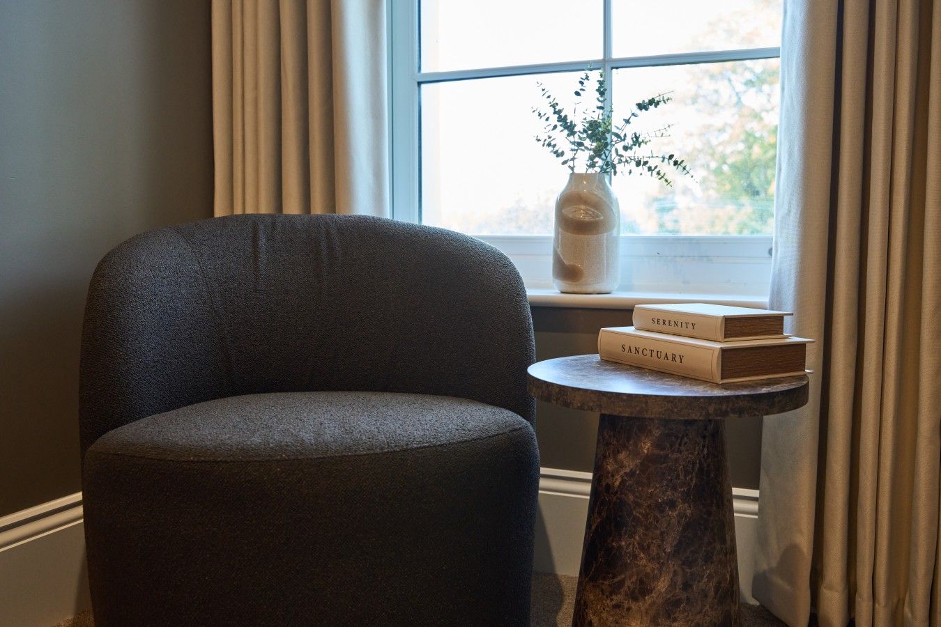 A dark grey armchair, a small round table with two books titled 'Serenity' and 'Sanctuary,' and a vase with a plant on a windowsill.