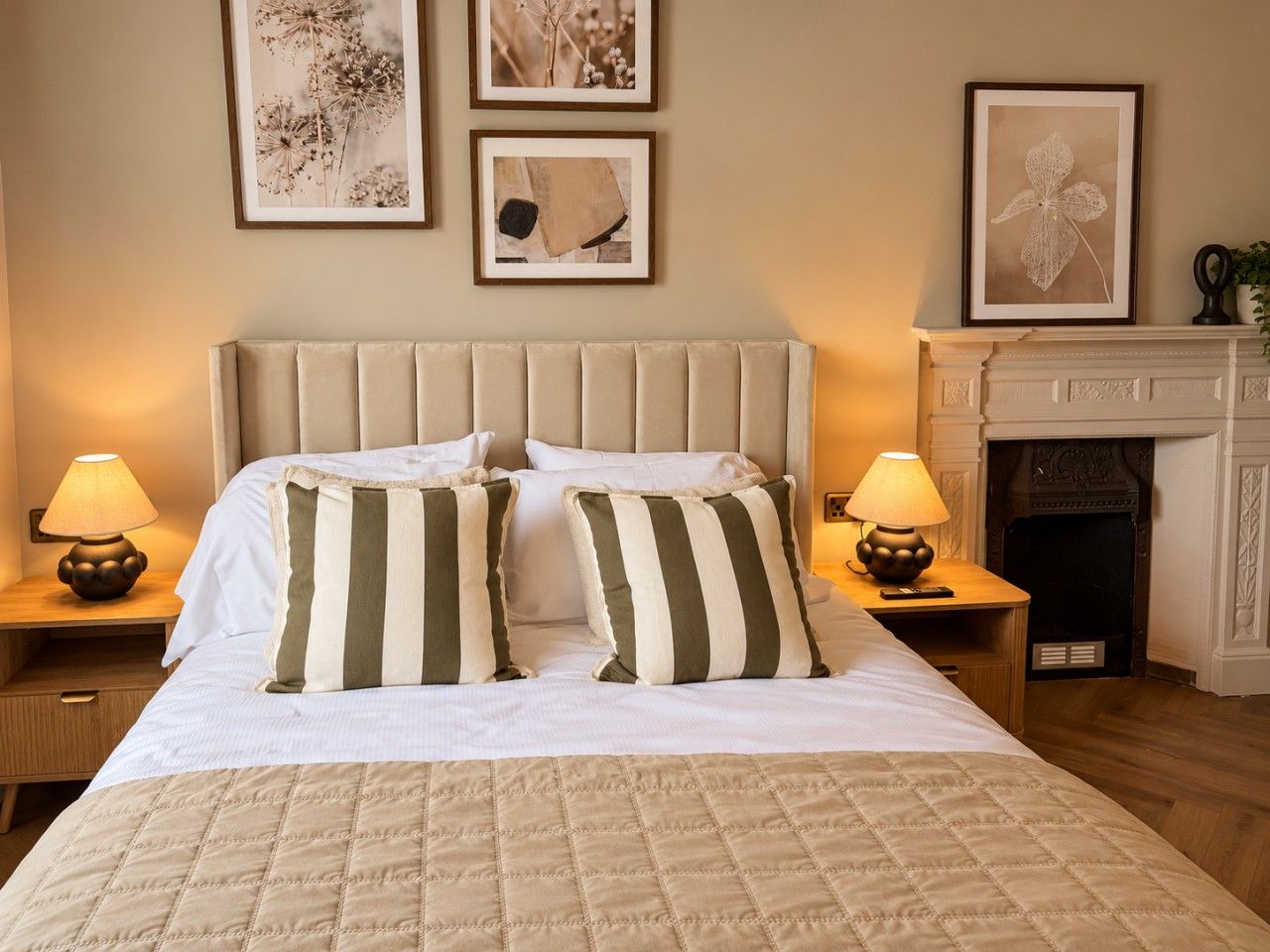 A hotel room with a large bed, two bedside lamps, a fireplace, and framed artwork on the wall.