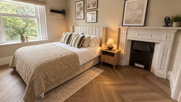 A bedroom with a bed, bedside table, lamp, fireplace, and framed pictures on the wall.