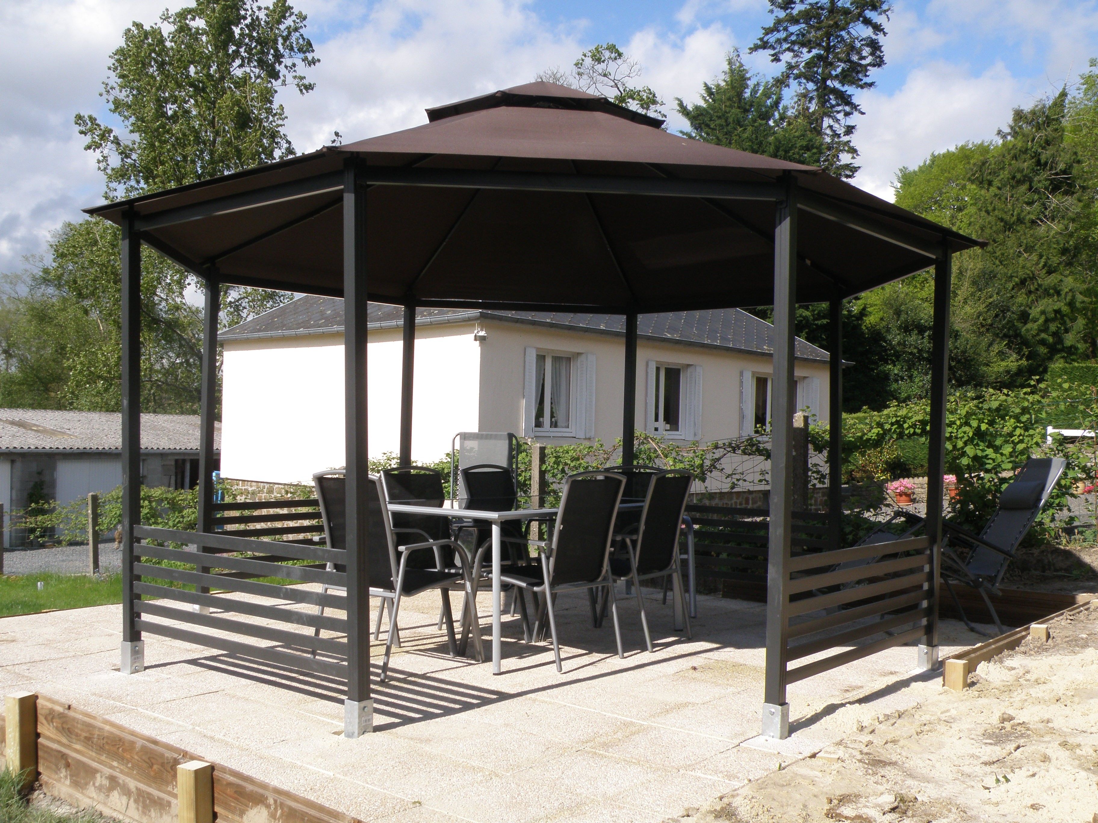 Un espace extérieur aménagé, abrité et doté de tables et de chaises, d’une structure en forme de pergola et d’où l’on peut admirer une maison en arrière-plan.