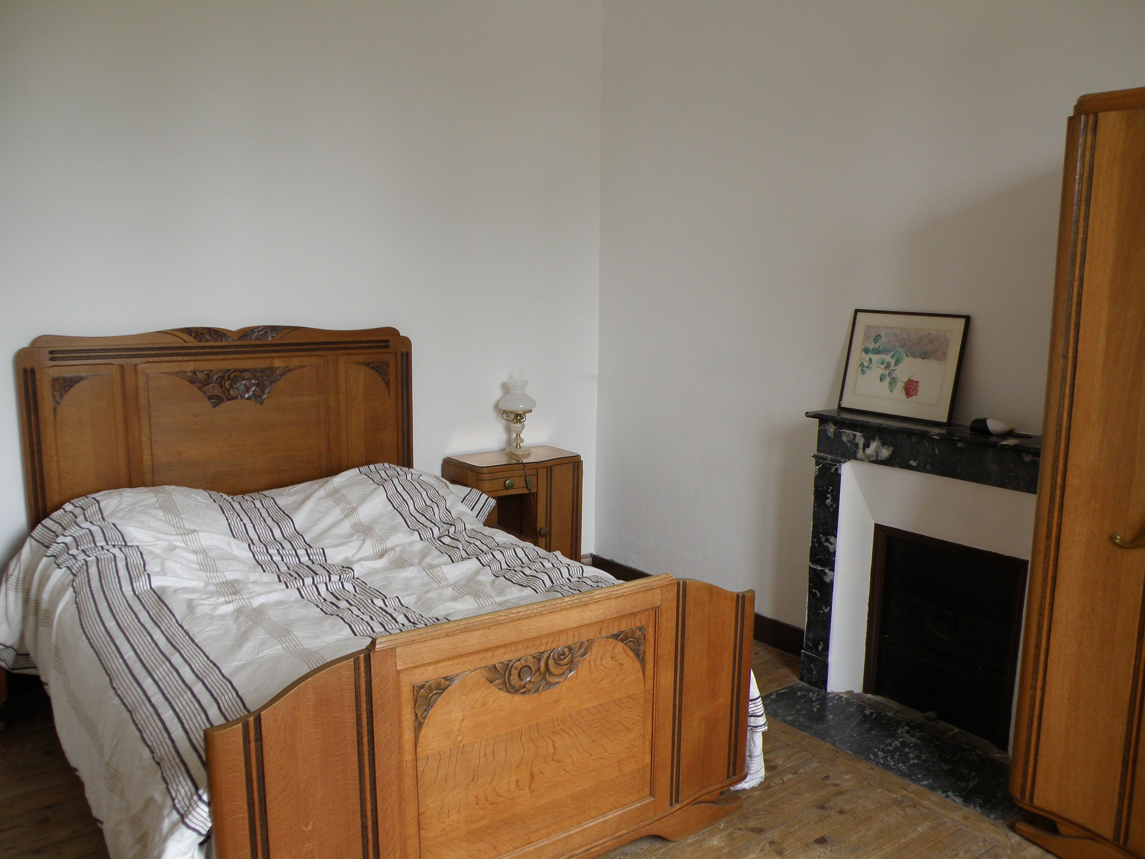 A bedroom with a wooden bed, a bedside table with a lamp, a fireplace, and a framed picture on the mantel.