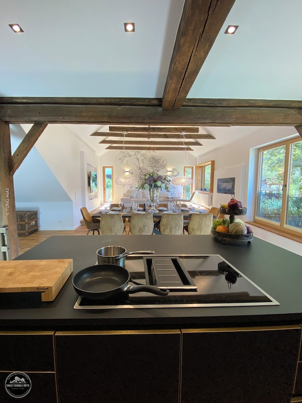 A modern kitchen with a central island, a dining area with a chandelier, and large windows.