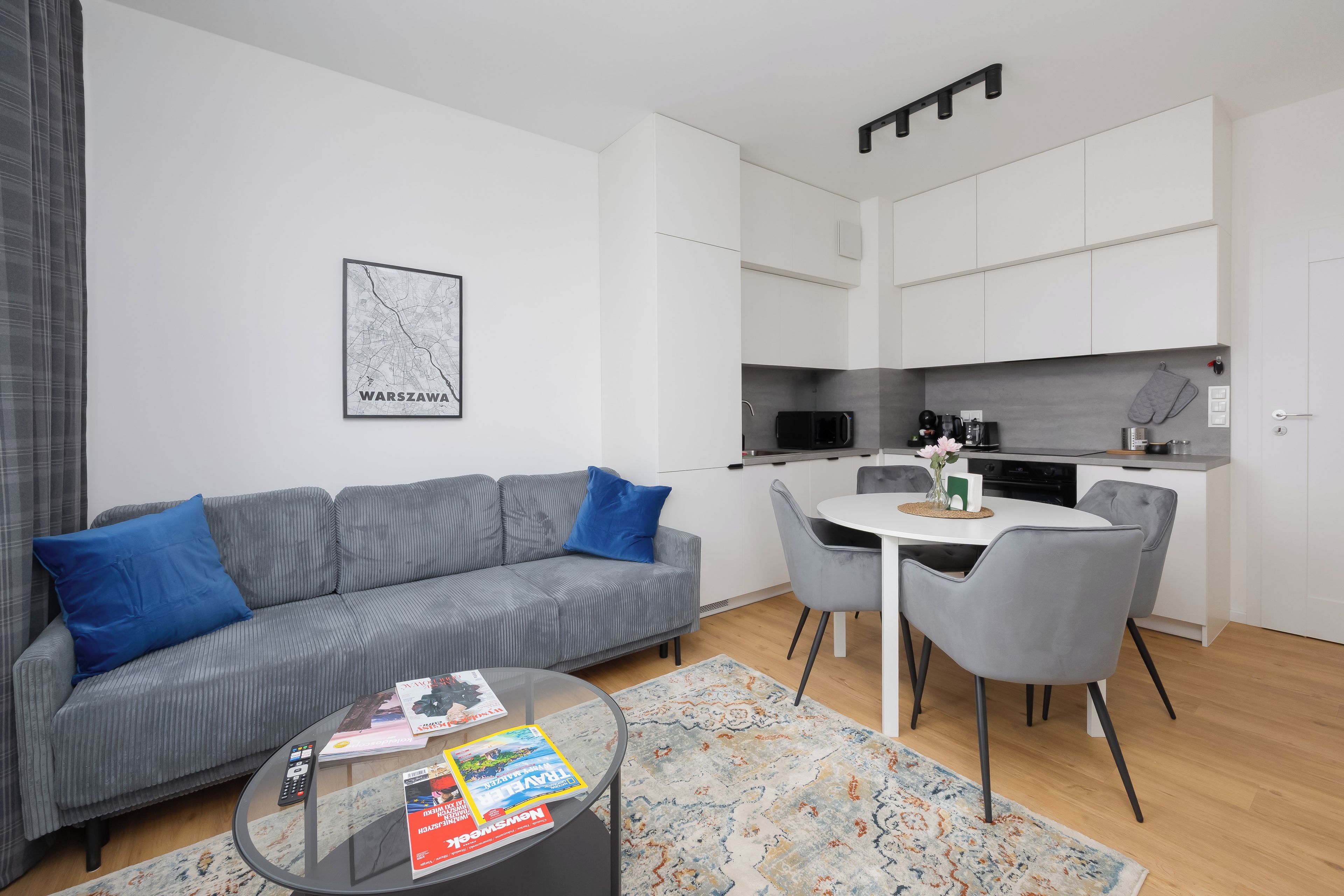 Een moderne woonkamer met een grijze bank, een salontafel met tijdschriften, en een keukenhoek met witte kasten en een magnetron.