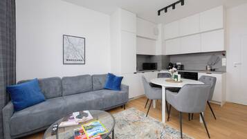 Een moderne woonkamer met een grijze bank, een salontafel met tijdschriften, en een keukenhoek met witte kasten en een magnetron.