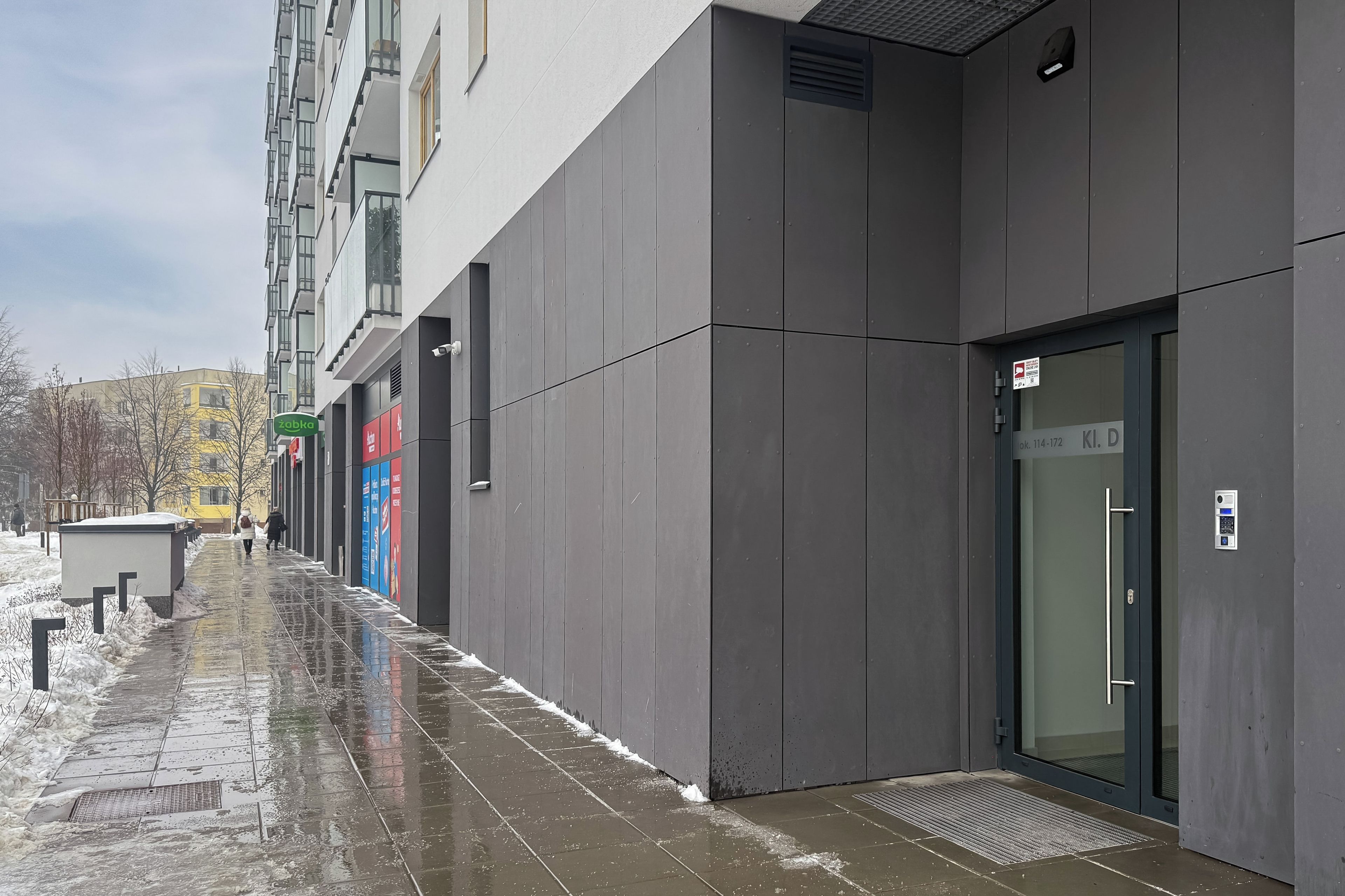A modern building with a glass entrance door, a visible address sign, and a wet pavement indicating recent rain.