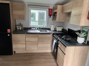 A modern kitchen with wooden cabinets, a black refrigerator, a stove, and a window with a view of the outside. - Harmony | Cosy 3BR Retreat in Windermere | Pet Friendly (Middleton)