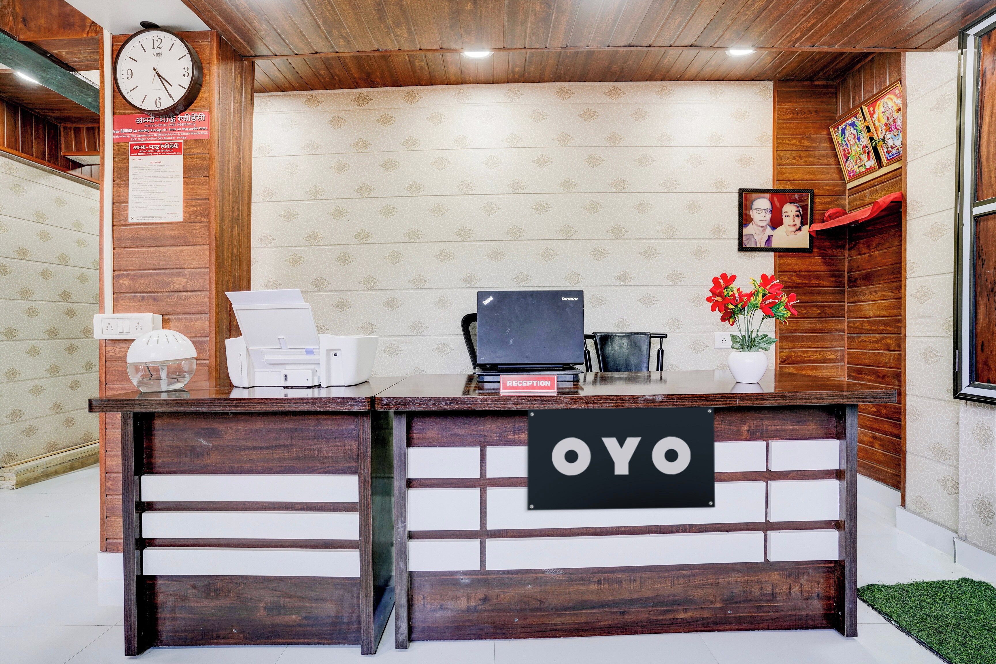 Un mostrador de recepción con un reloj, una computadora portátil Lenovo, una impresora y un florero con flores.