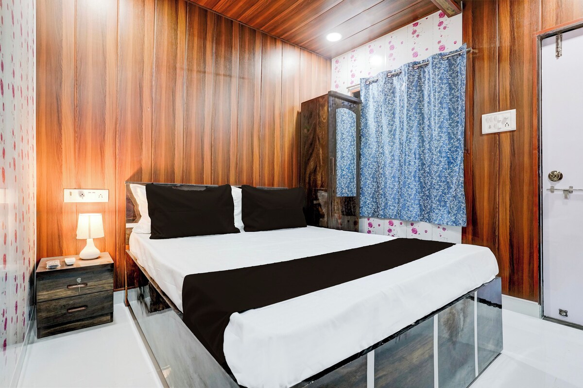 Un dormitorio con pared de madera, una cama con ropa de cama blanco y negro, una mesita de noche con una lámpara y un baño con cortina de ducha.
